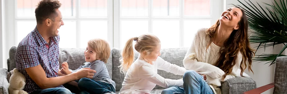 Man, woman and two kids all sitting on a grey couch, laughing and tickling each other